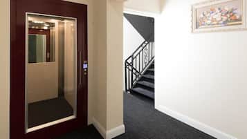 Un ascenseur doté d’un écran numérique, un escalier avec une rampe noire et une œuvre d’art florale encadrée accrochée au mur.