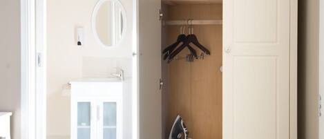 A room with a white cabinet, a mirror, and a wooden wardrobe with hangers and an iron.