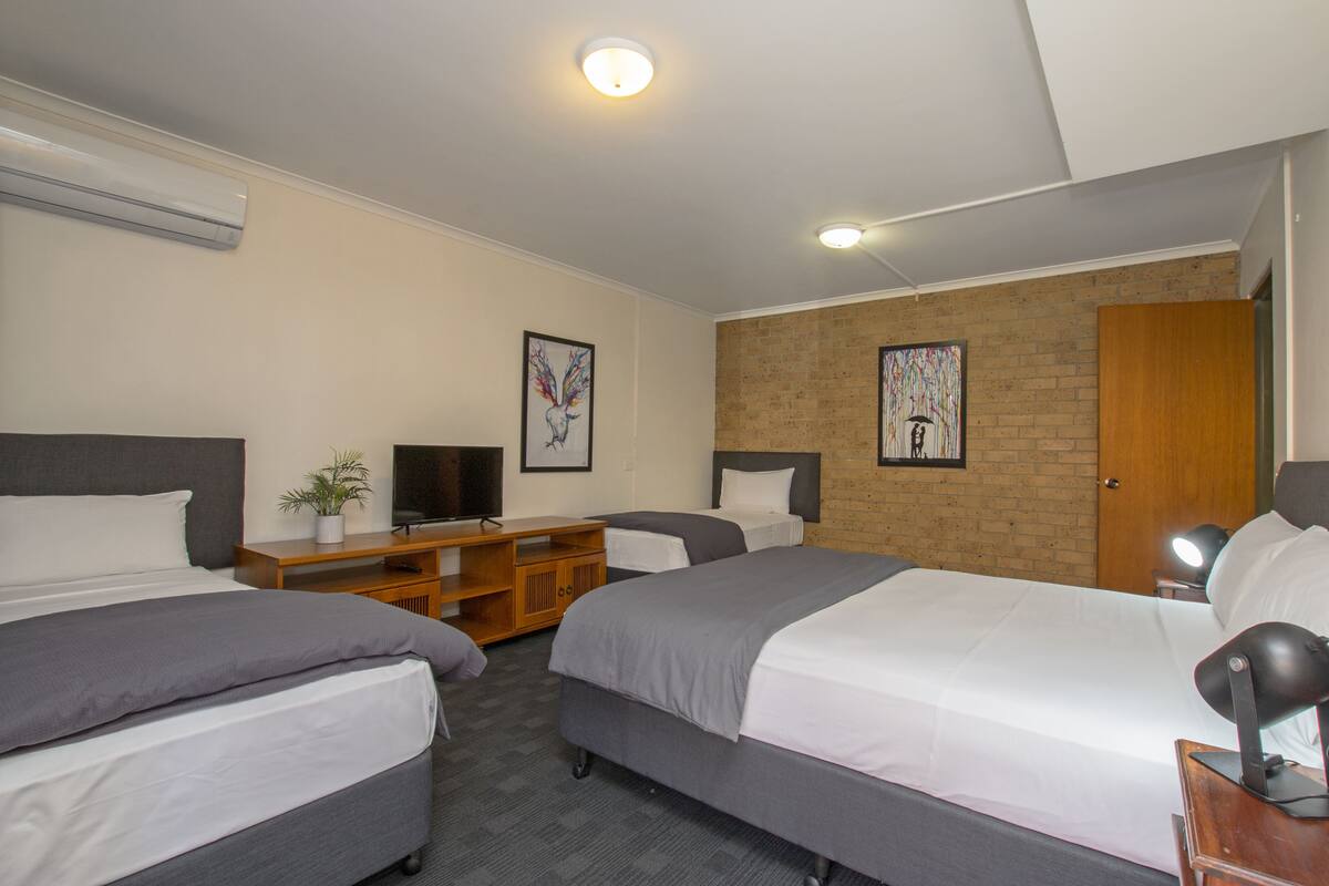 A hotel room with two beds, a TV, a desk, and a brick wall.