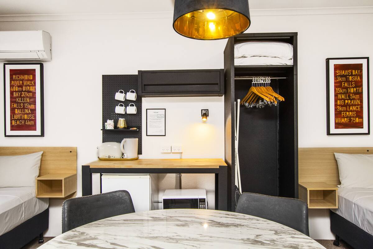 A hotel room with a bed, a desk, a chair, a table, a microwave, a hanging light fixture, and a wardrobe.