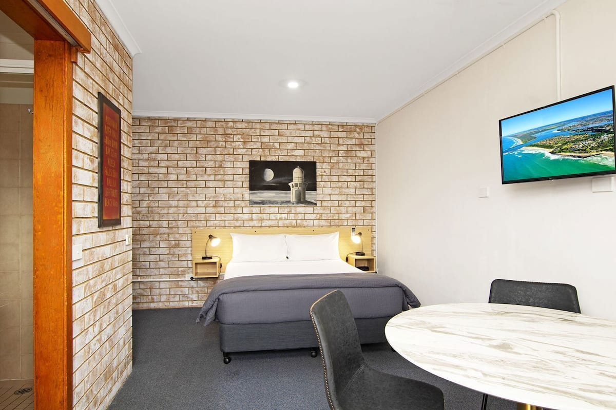 A hotel room with a brick wall, a bed with a headboard, bedside lamps, a flat-screen TV, a round marble table, and two chairs.
