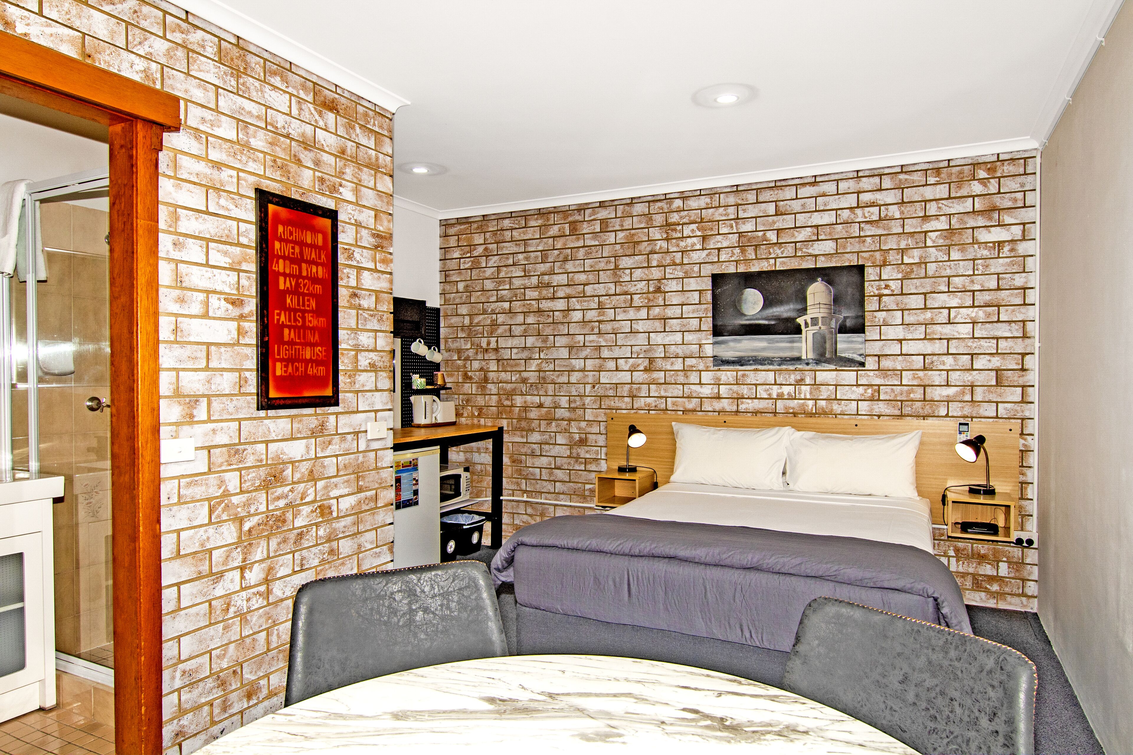 A hotel room with a brick-patterned wall, a bed with a wooden headboard, a nightstand, a desk with a lamp, and a framed picture on the wall.