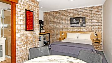 A hotel room with a brick-patterned wall, a bed with a wooden headboard, a nightstand, a desk with a lamp, and a framed picture on the wall.