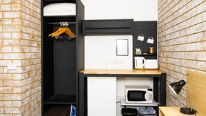Una habitación de hotel compacta con armario, pequeña cocina, escritorio con lámpara y pared con diseño de ladrillo.