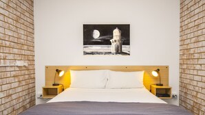 Una cama bien hecha con un edredón gris, dos mesitas de noche con lámparas y un cuadro colgado en la pared de un faro.