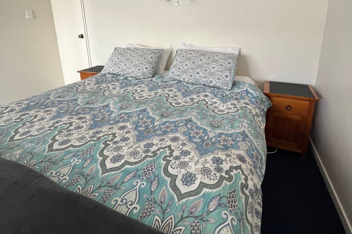 Une chambre à coucher avec un lit, une literie à motifs, une table de chevet, un tiroir en bois et un luminaire fixé au mur.