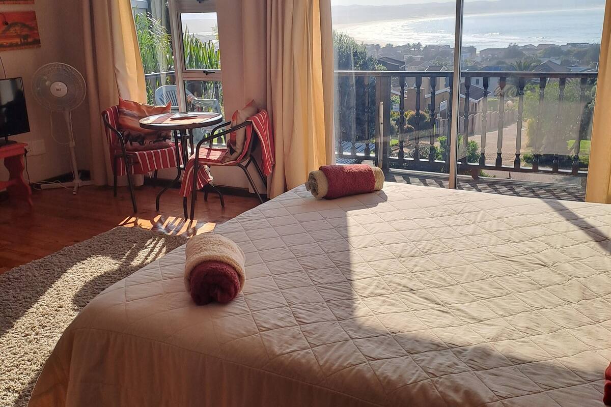 A bedroom with a large bed, a small table, and a chair. There is a balcony with a view of the ocean.