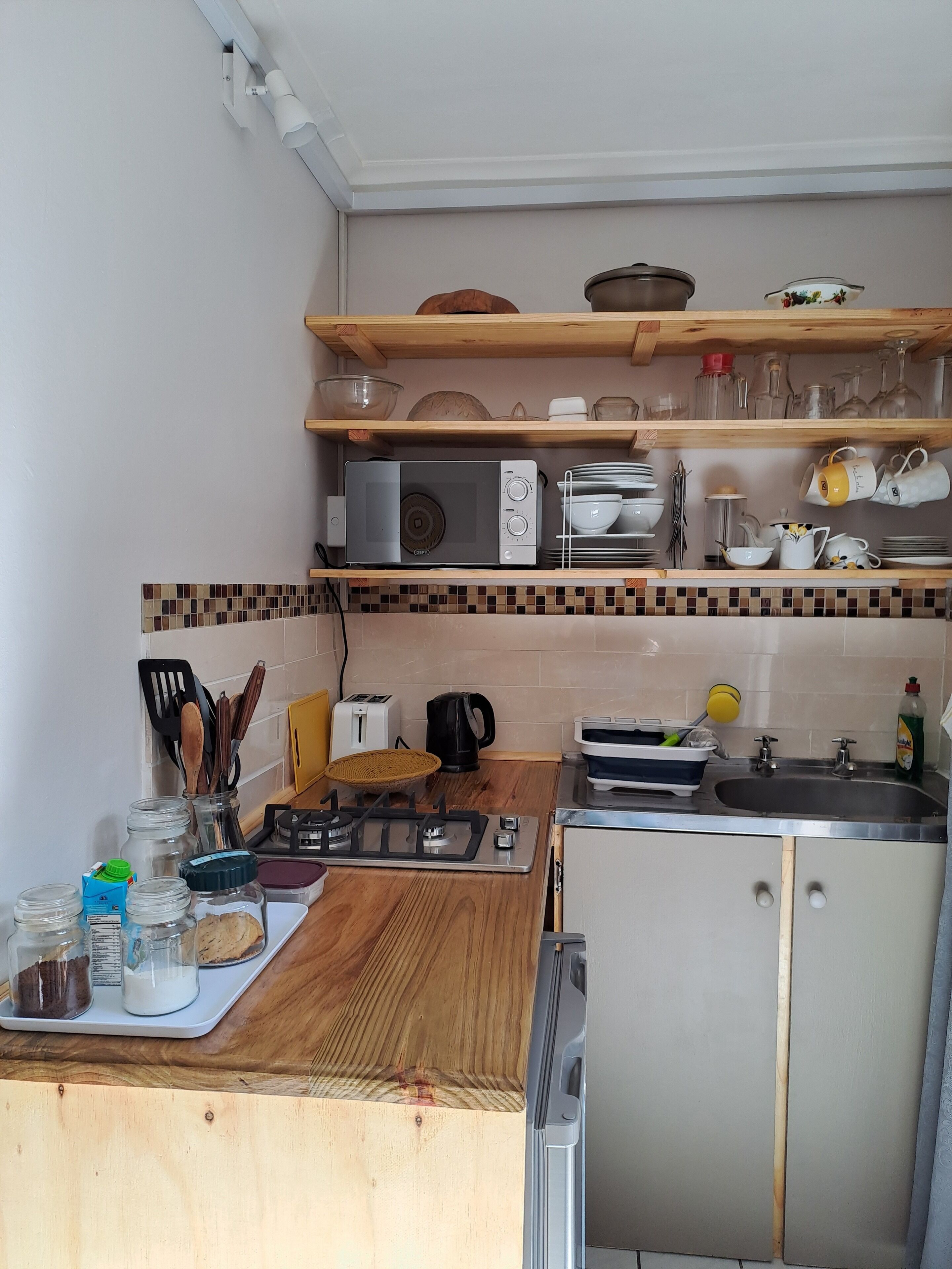 Una cucina con un piano di lavoro in legno, una stufa a gas, un lavello e un forno a microonde. Sopra il piano di lavoro ci sono scaffali con diversi oggetti da cucina.