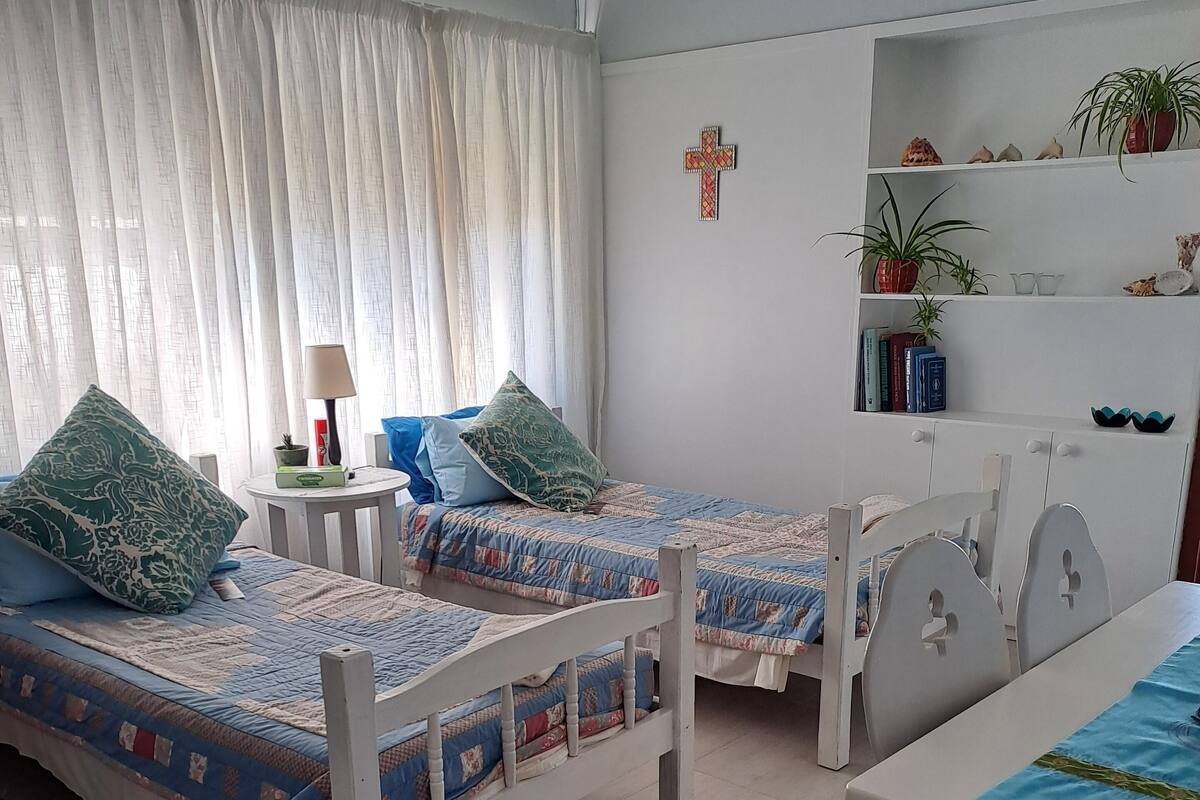A bedroom with two beds, a bedside table with a lamp, a shelf with decorative items, and a window with curtains.