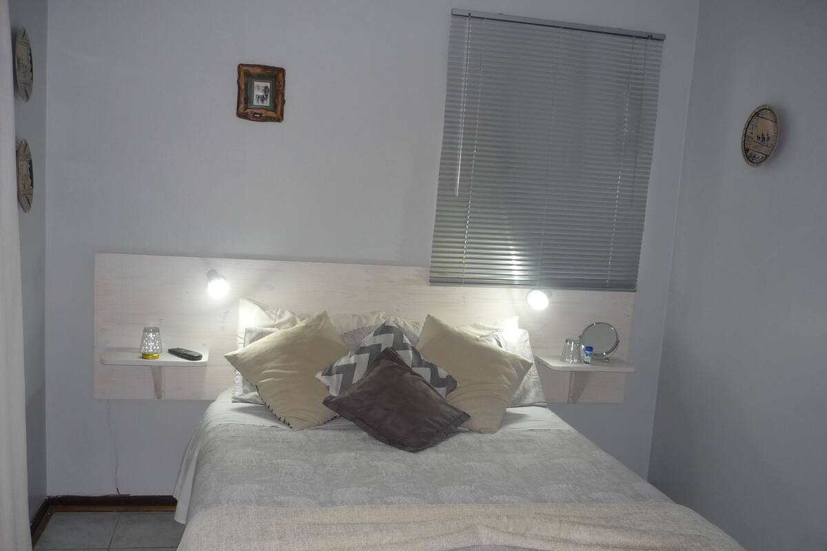 A neatly made bed with a headboard, bedside tables with lamps, and a window with blinds.