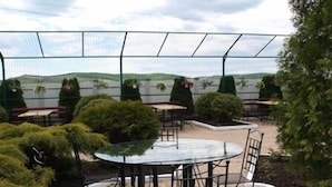A covered outdoor seating area with tables and chairs, surrounded by greenery and a view of the landscape.
