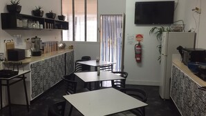 A small, compact kitchen with basic amenities including a coffee maker, a refrigerator, and a fire extinguisher. There are several tables and chairs for dining, and the kitchen features a window with blinds. - Maryborough Motel and Conference Centre (Maryborough)