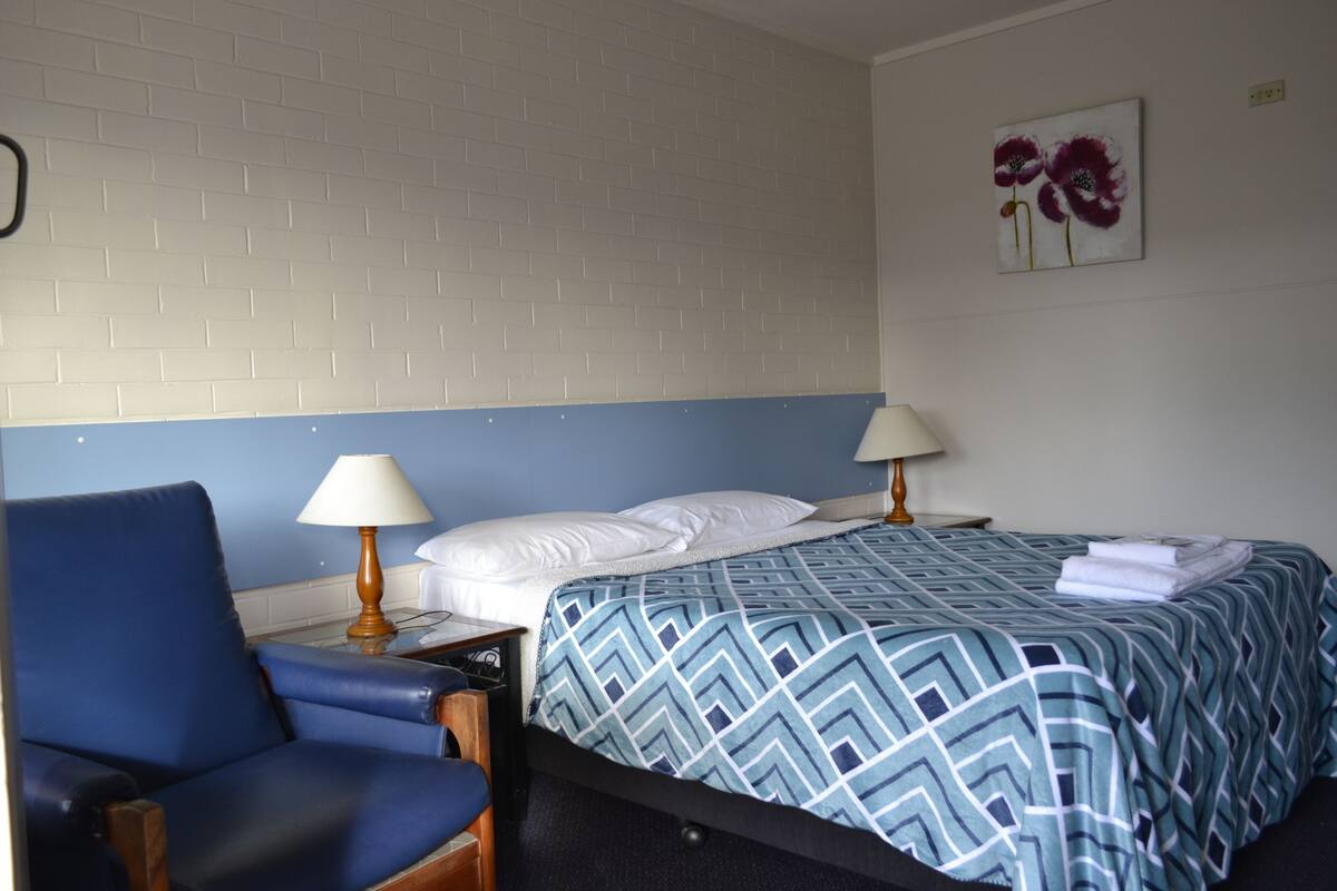 Ein Hotelzimmer mit einem Einzelbett, einem blauen Sessel, zwei Nachttischlampen und einem Wandbild mit Blumen.