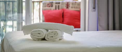 A neatly made bed with rolled white towels and a view of a balcony with a red cushion.