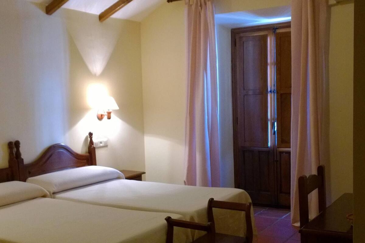 Une chambre d’hôtel avec deux lits, une petite table, une chaise, une lampe et une fenêtre avec des rideaux.