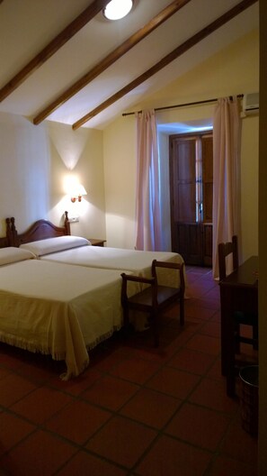 A hotel room with two beds, a small table, a chair, a lamp, and a window with curtains. - Hotel Palacete de Mañara (Montejaque)
