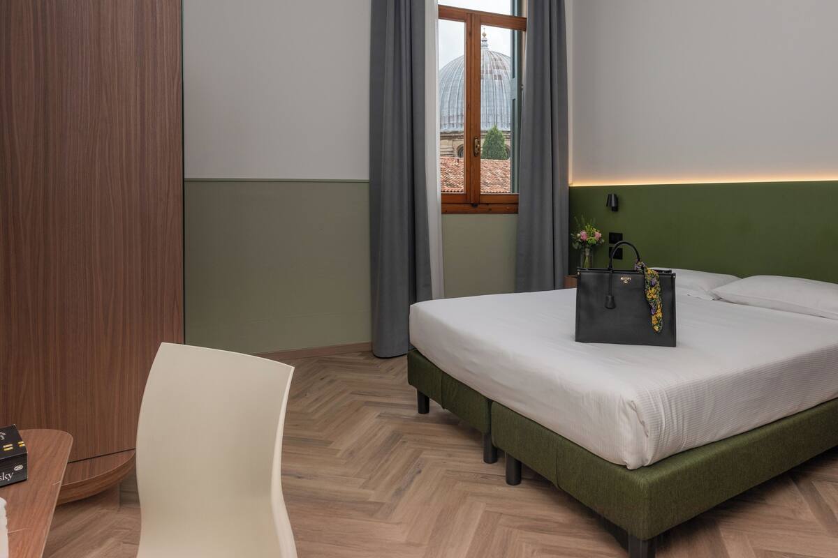Habitación de hotel con cama, una silla, una mesita de noche, lámpara y vista a una cúpula a través de una ventana.