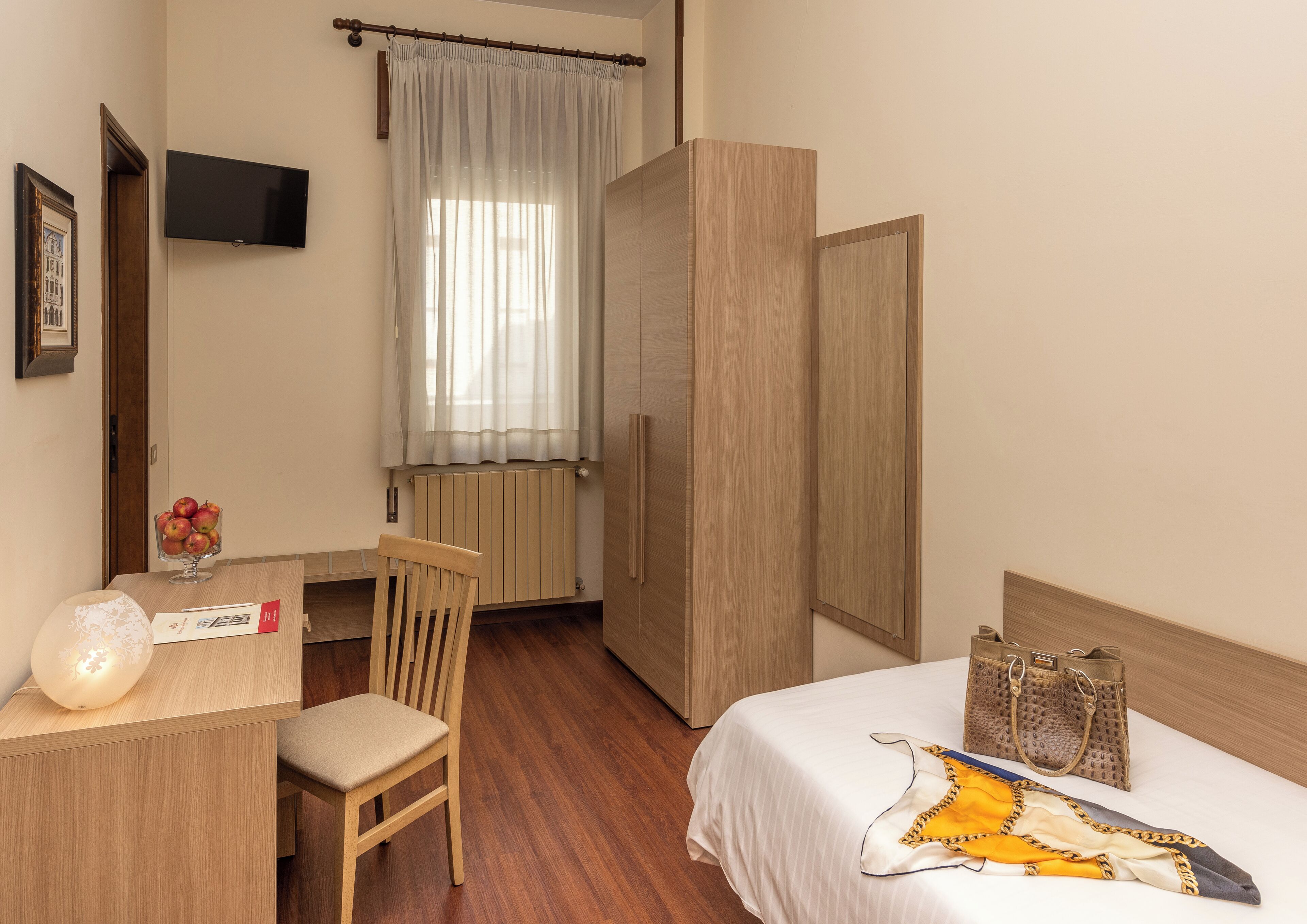 A hotel room with a wooden desk, chair, bed with a blanket, a radiator, a TV mounted on the wall, and a wardrobe.