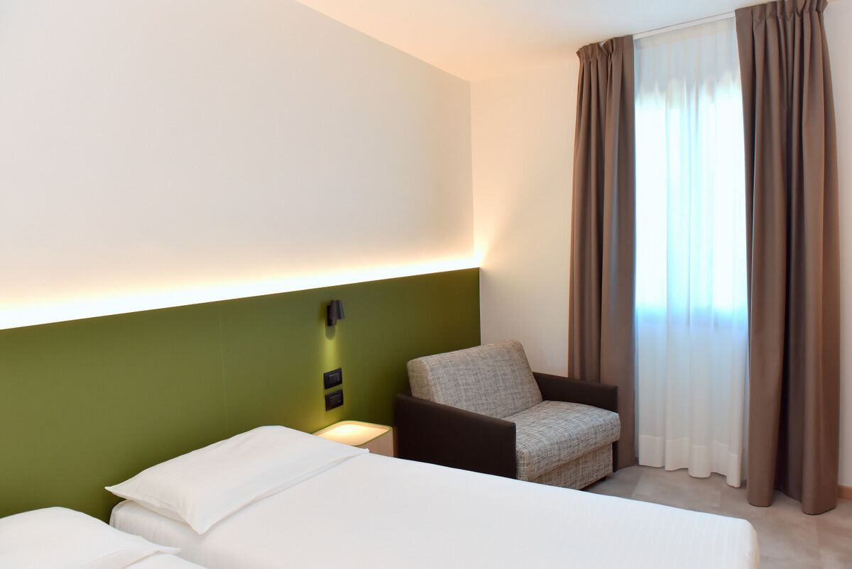 A hotel room with a green accent wall, a bed, a chair, a nightstand, and a window with curtains.