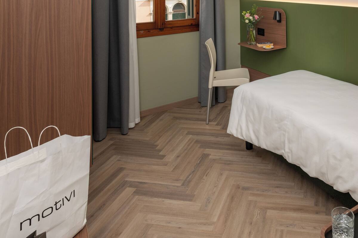 Habitación de hotel con cama, televisor montado en la pared, armario de madera, ventana con cortinas y una mesita con un jarrón y un control remoto.