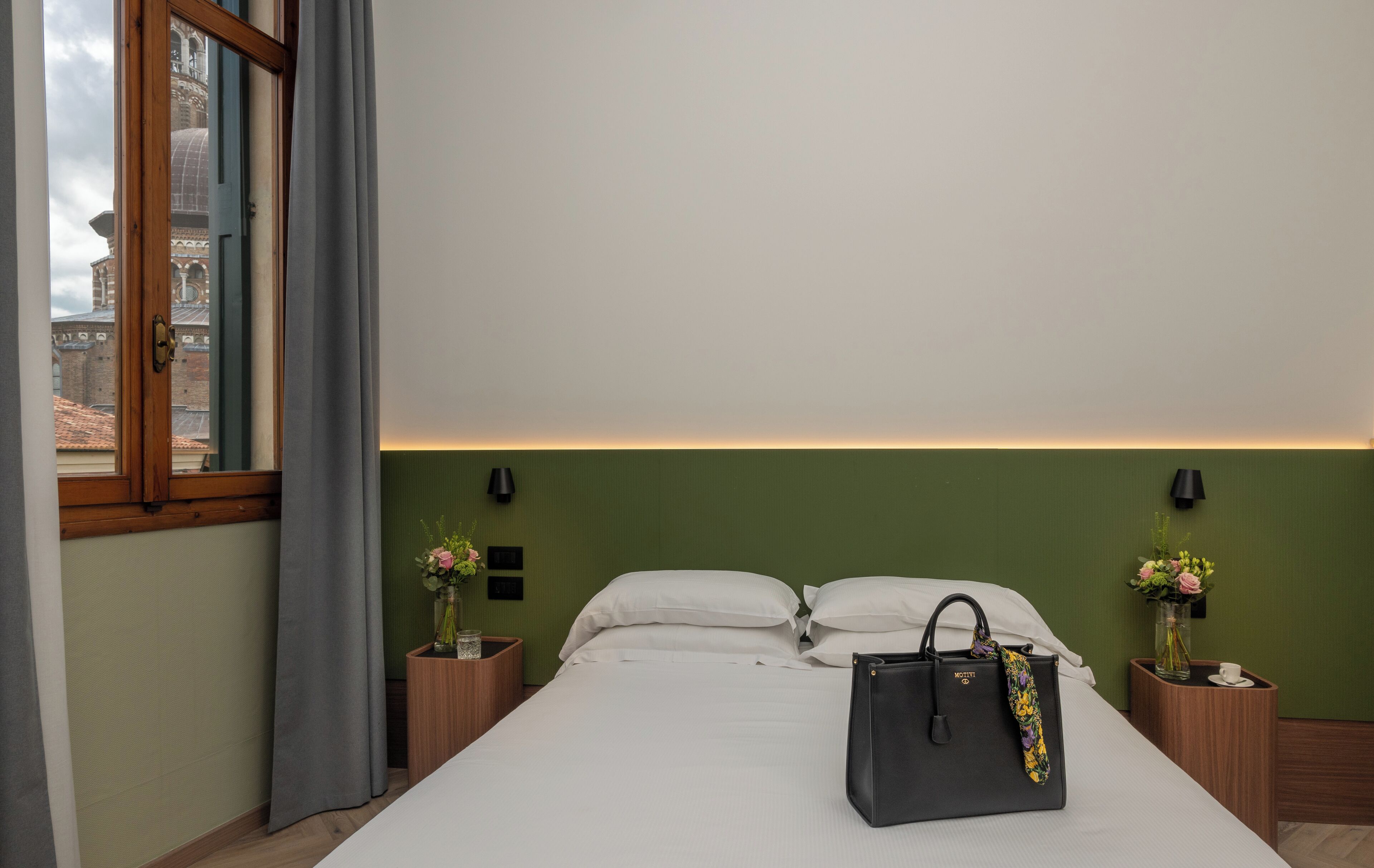 A hotel room with a bed, a nightstand with a vase of flowers, a wooden nightstand with a lamp, and a view of a building through a window.