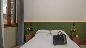 A hotel room with a bed, a nightstand with a vase of flowers, a wooden nightstand with a lamp, and a view of a building through a window.