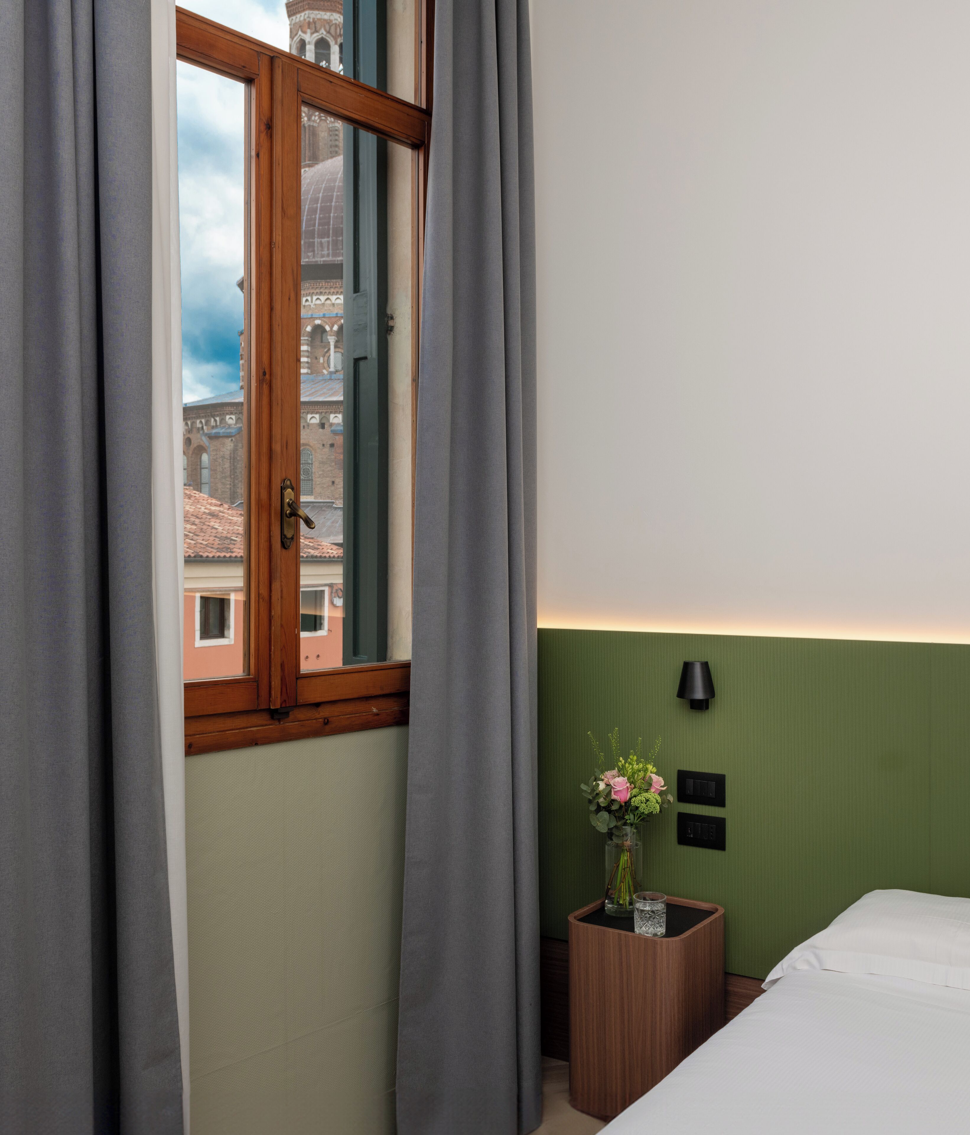 A hotel room with a view of a historic building through a window with wooden frames and grey curtains.