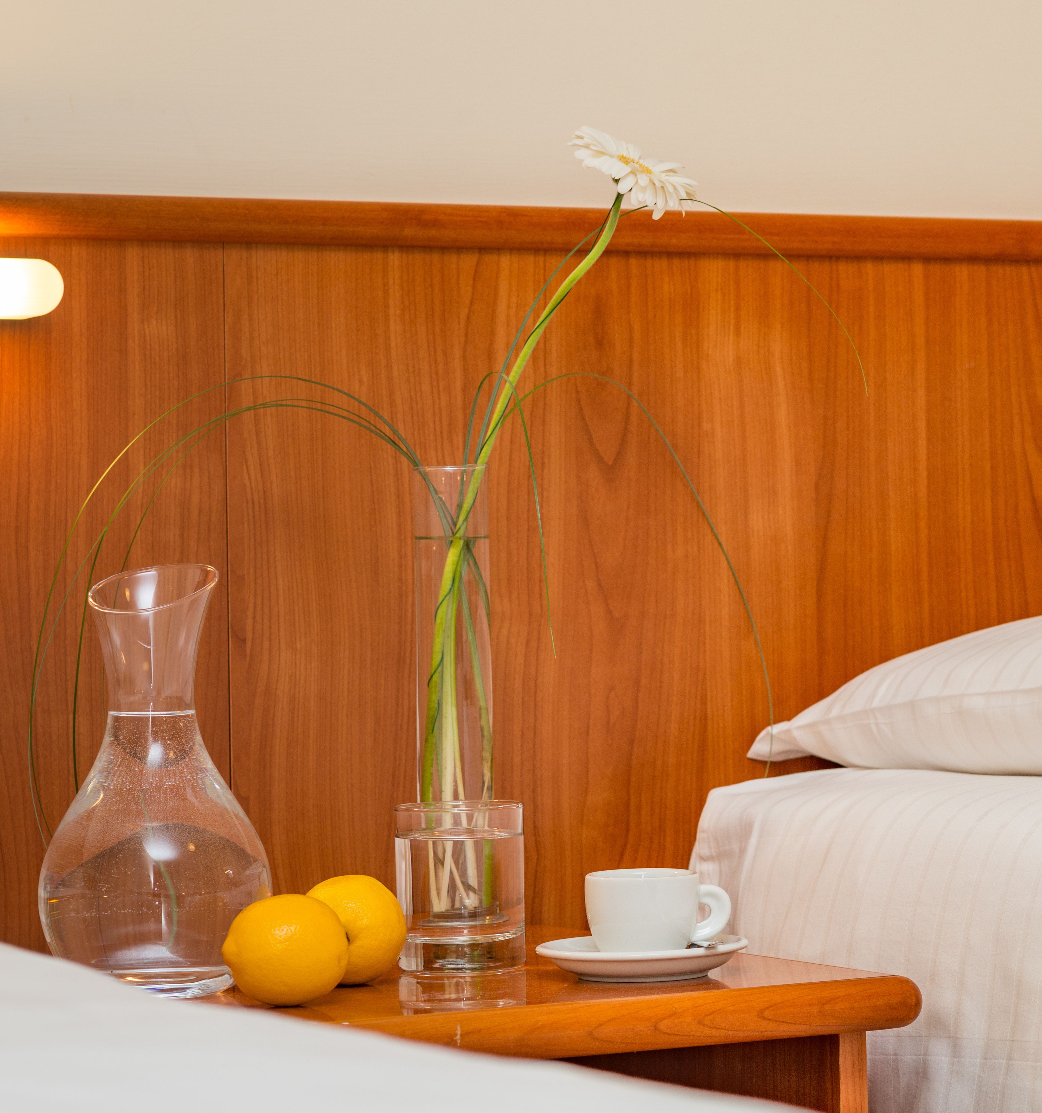 A wooden bedside table with a clear vase containing a single white flower, a glass of water, and two lemons.