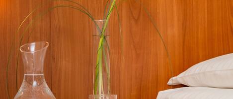 Una mesita de noche de madera con un florero transparente que contiene una flor blanca, un vaso de agua y dos limones.