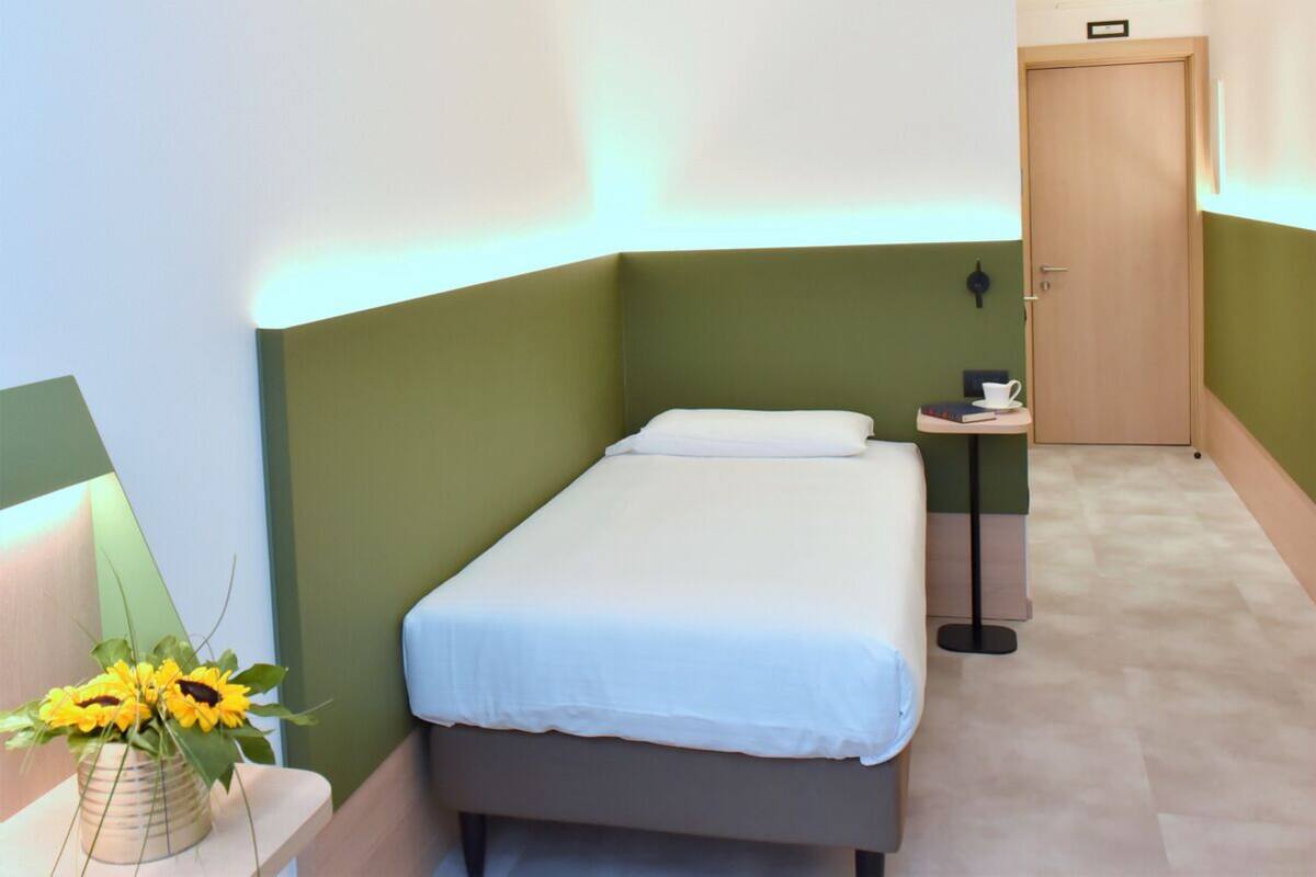 Una habitación pequeña y ordenada con una cama individual, una mesita de noche con una taza y un libro, y una puerta.