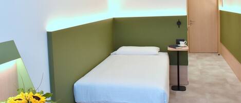 Una habitación pequeña y ordenada con una cama individual, una mesita de noche con una taza y un libro, y una puerta.