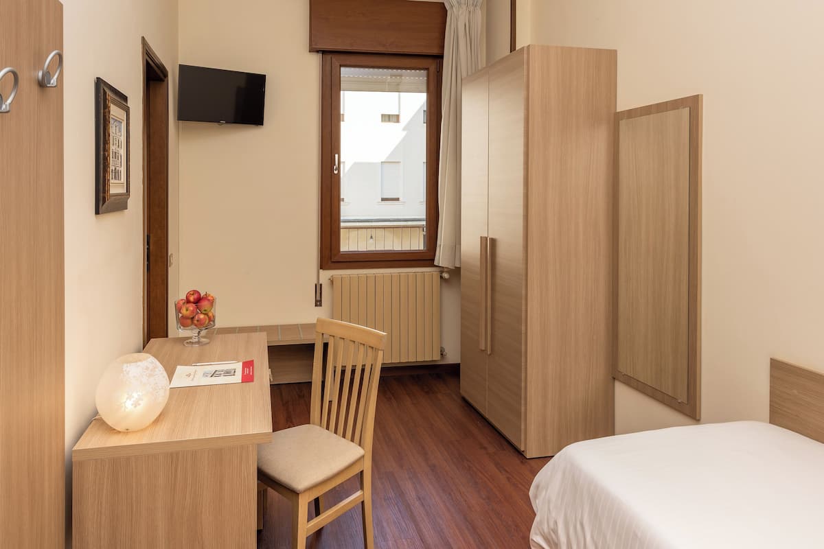 Una habitación de hotel con una cama, un escritorio con silla, un televisor en la pared, una ventana con cortinas y un radiador.