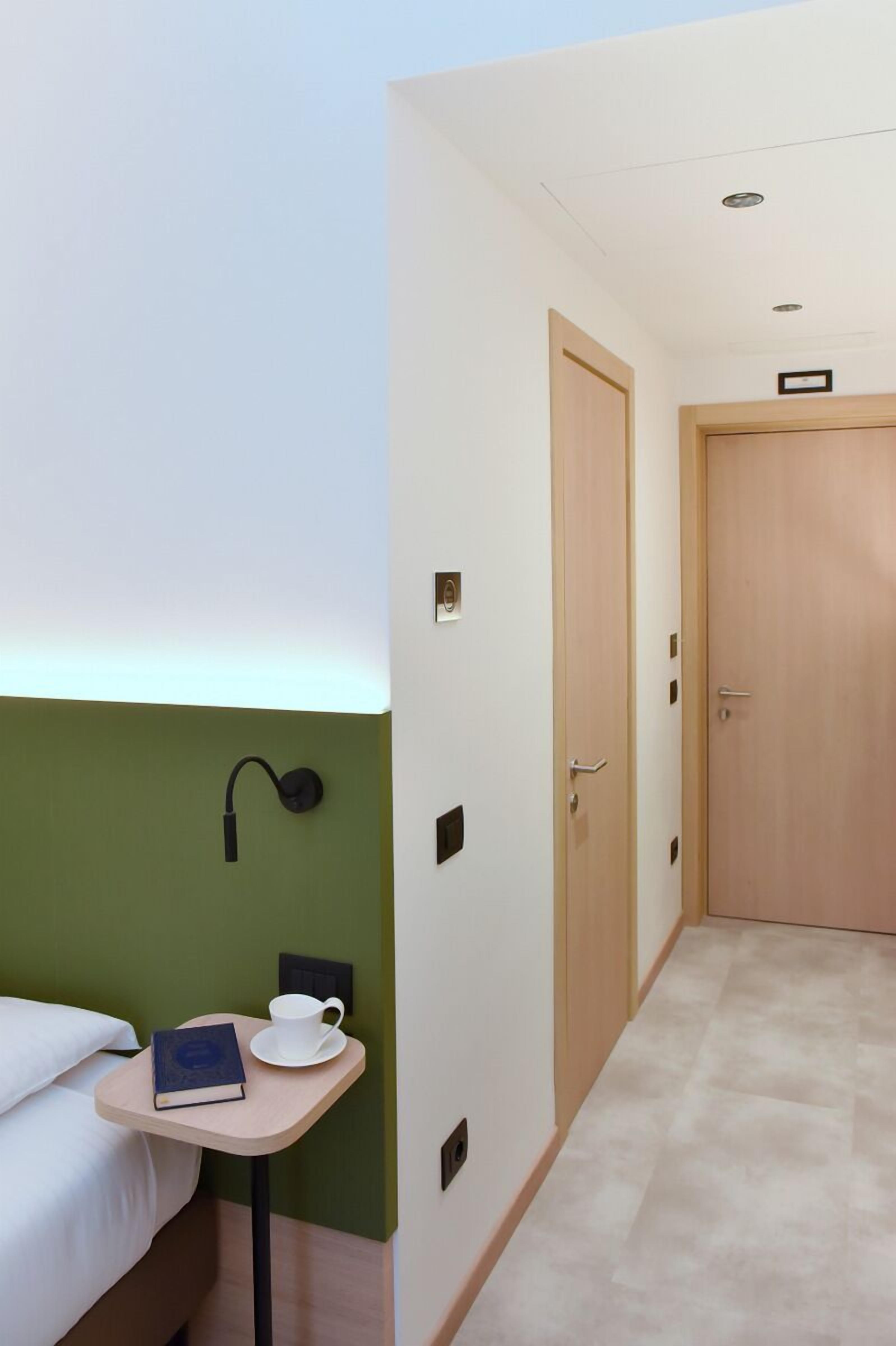 Une chambre d’hôtel avec un lit, une petite table sur laquelle se trouvent un livre et une tasse, une lampe fixée au mur et une porte donnant sur une autre pièce.
