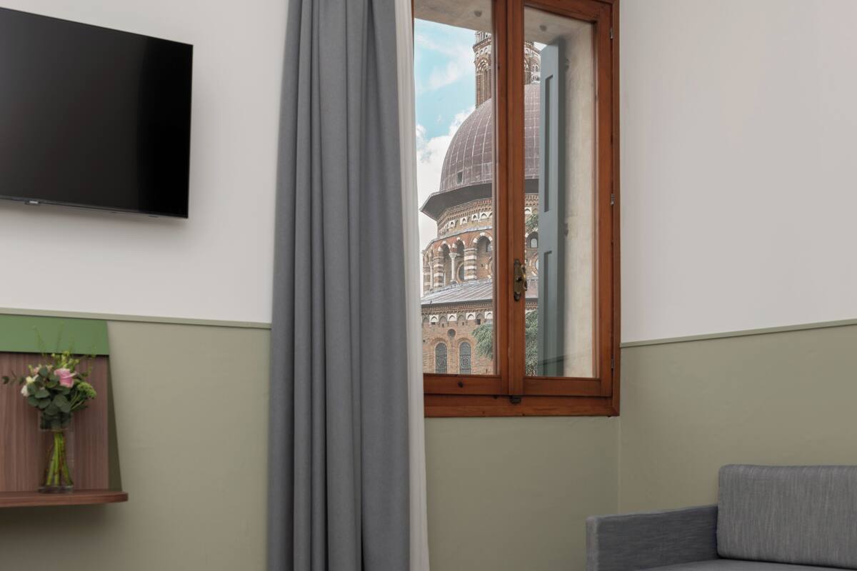 Una habitación con televisión en la pared, ventana con vista a un edificio histórico y un sofá gris.