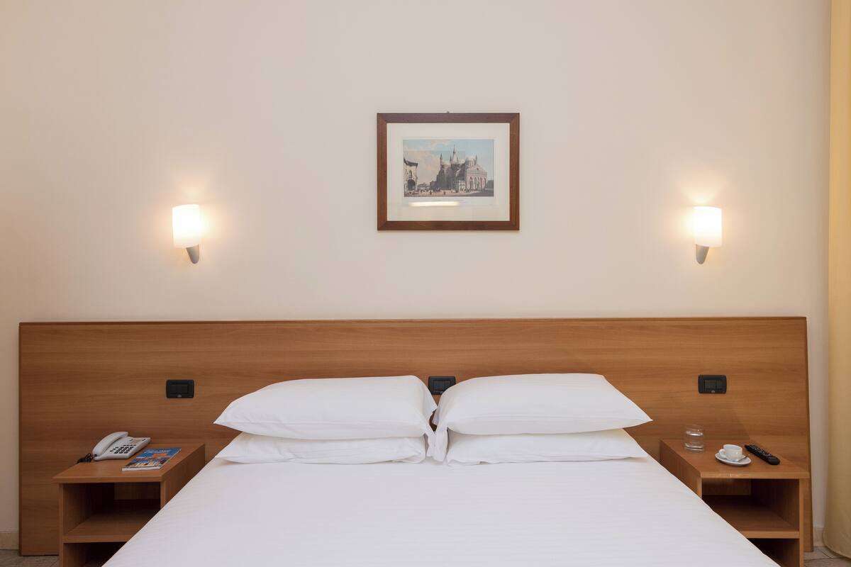 Una cama bien hecha con ropa blanca, dos mesitas de noche con un teléfono y un control remoto, y un cuadro en la pared.