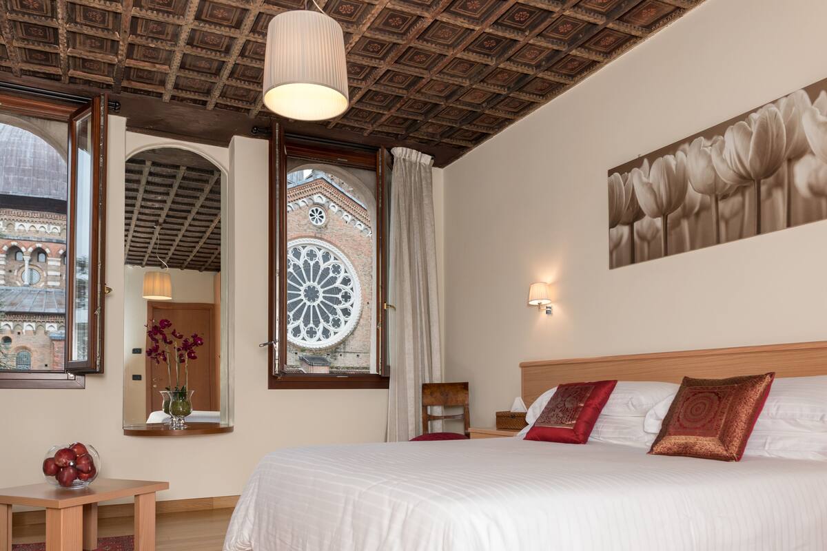 Una habitación de hotel con una cama grande, cabecera de madera, candelabro, ventana amplia con vista a un edificio y un cuadro colgado en la pared.