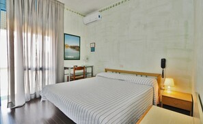 A hotel room with a large bed, a desk, a chair, a nightstand with a lamp, and an air conditioning unit on the wall. - Hotel Antonella (Caorle)