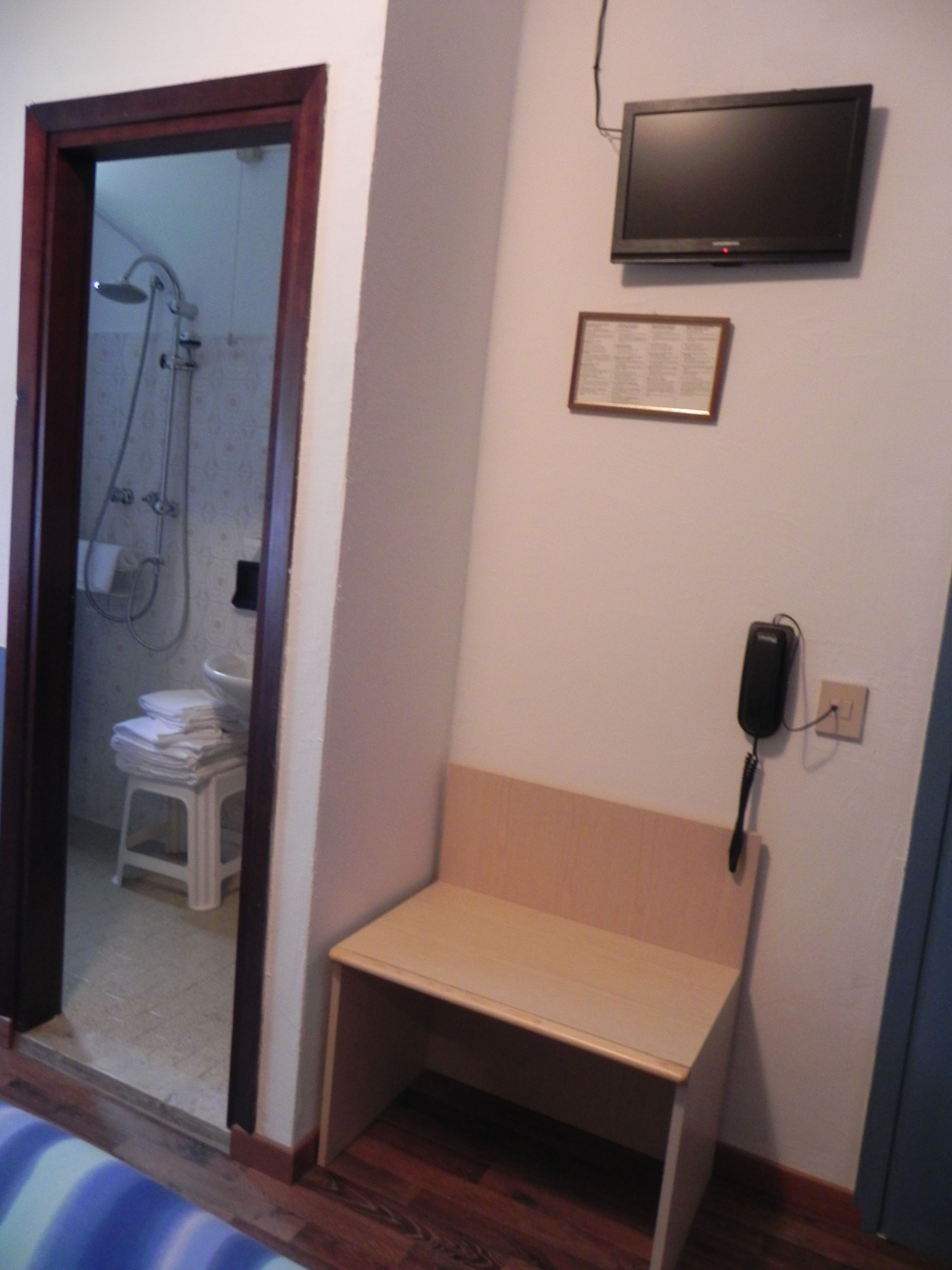 Een badkamer met een douche, toilet, wastafel, een klein bankje, een ingelijst certificaat en een telefoon.