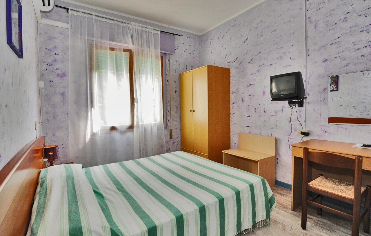 A hotel room with a bed, a wooden wardrobe, a desk, a chair, a television, and a window with curtains.