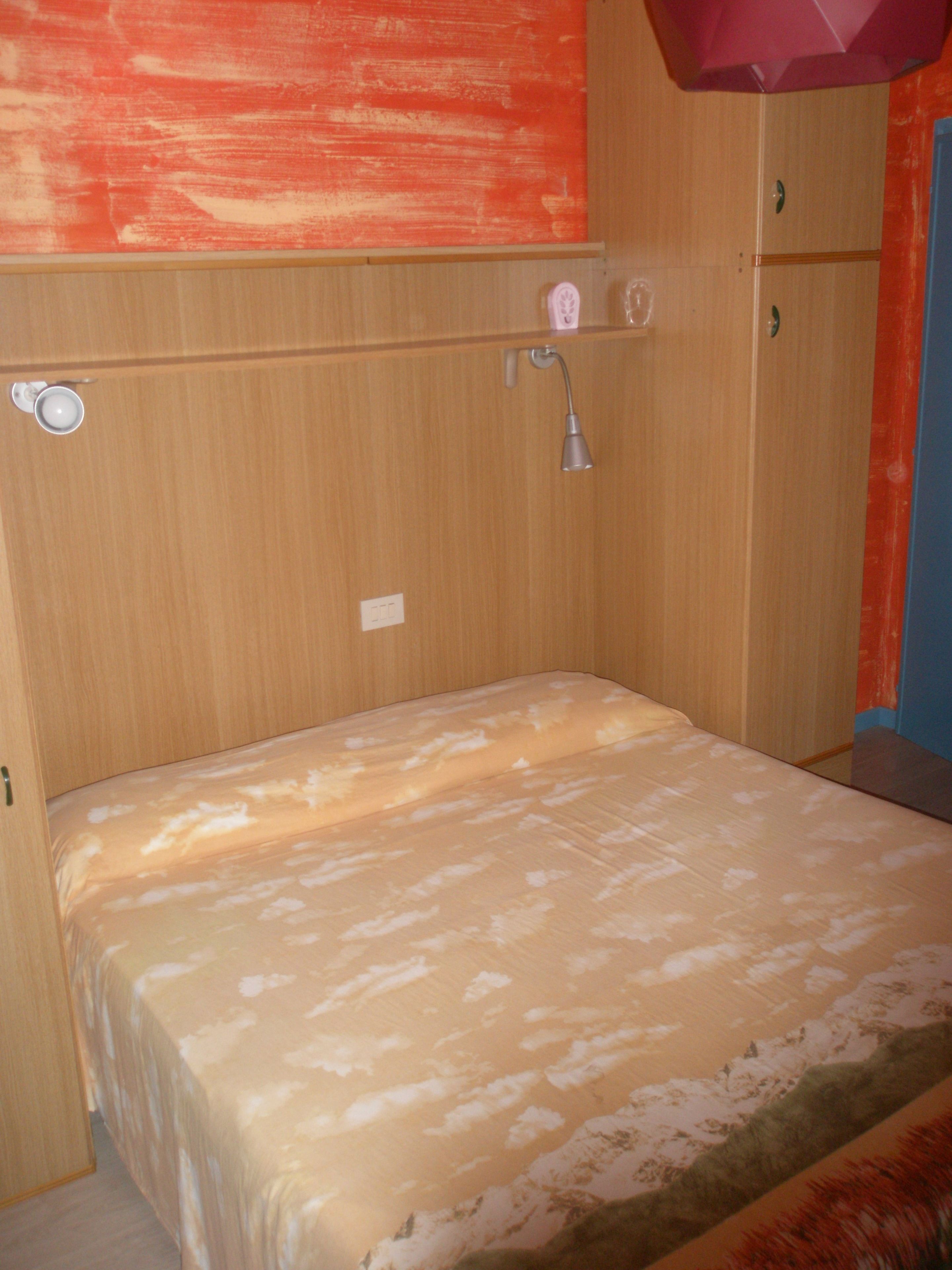 A small room with a single bed, a wooden wardrobe, a small shelf with toiletries, and a shower area.