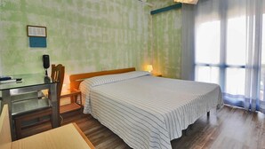 A room with a bed, a desk, and a chair. There is a window with curtains, a wall-mounted air conditioner, and a lamp on the desk. - Hotel Antonella (Caorle)