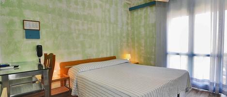 A room with a bed, a desk, and a chair. There is a window with curtains, a wall-mounted air conditioner, and a lamp on the desk.