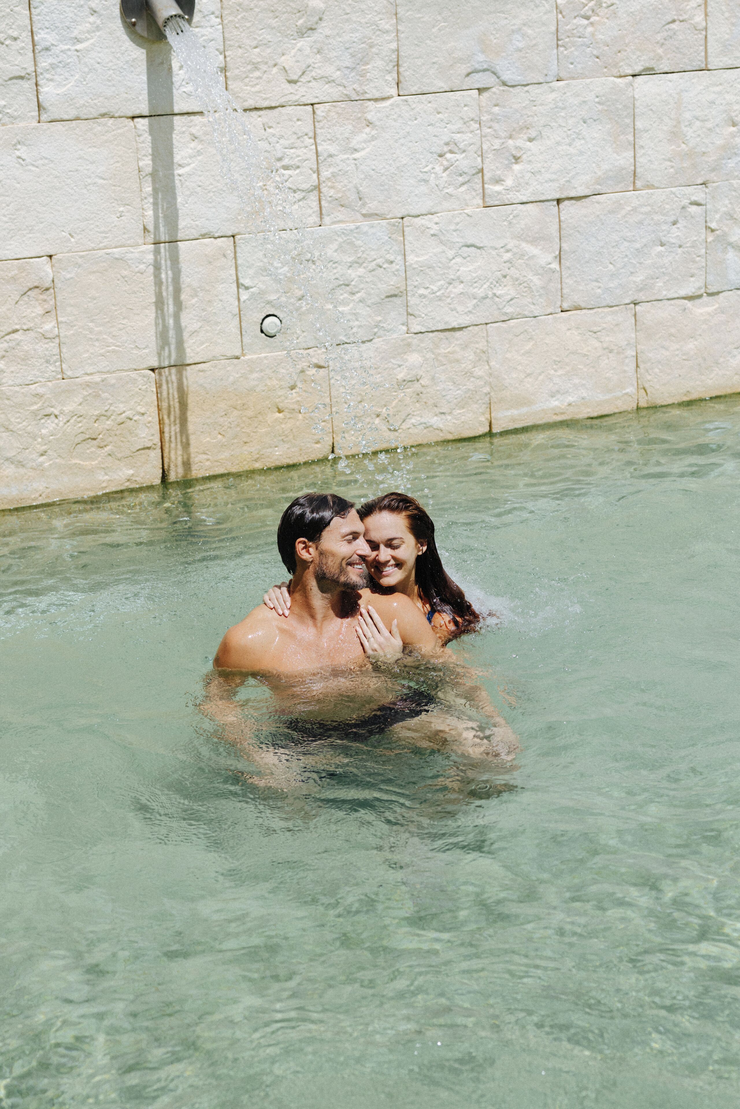 Un hombre y una mujer en una piscina poco profunda con un muro de piedra al fondo.