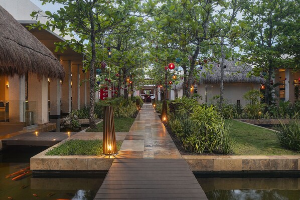 Un camino conduce a una piscina con edificios de techo de paja, rodeado de vegetación e iluminación decorativa.