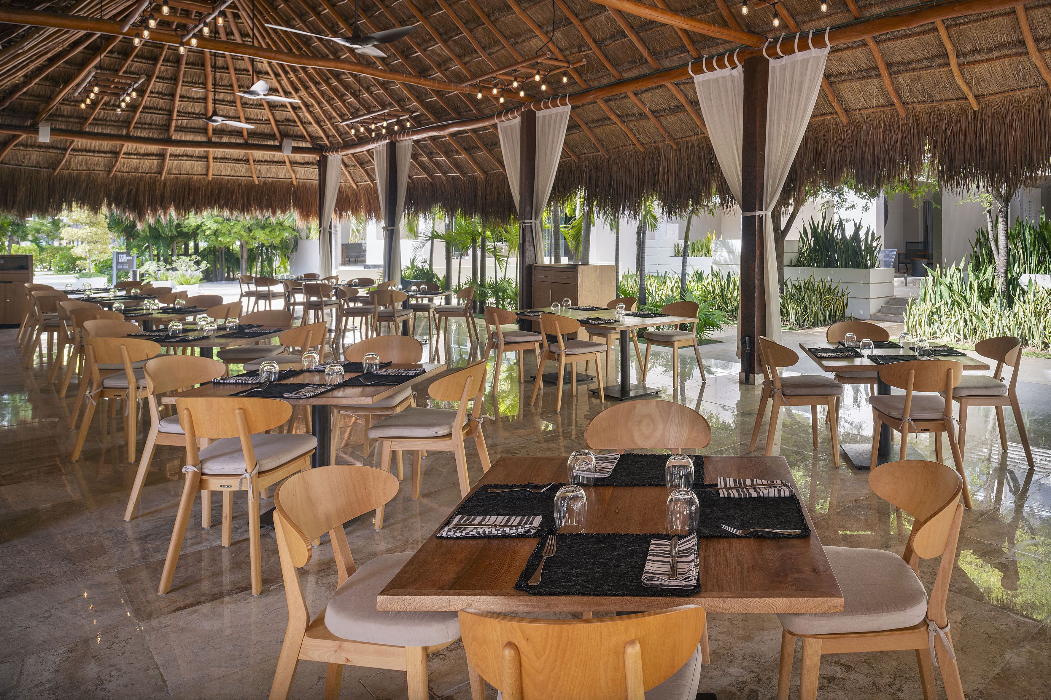 Un área de comedor al aire libre con mesas y sillas de madera, techo de paja y plantas en macetas.