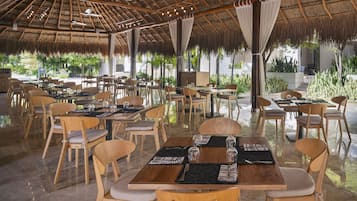 Un área de comedor al aire libre con mesas y sillas de madera, techo de paja y plantas en macetas.