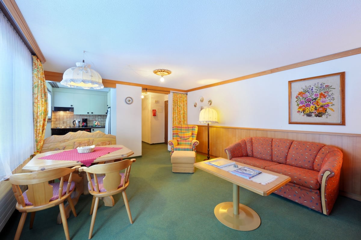 Una sala de estar compacta con una mesa de comedor, sillas, un sofá, un sillón pequeño, una lámpara y un cuadro en la pared.