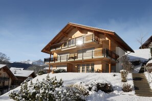 A two-story wooden chalet with a balcony, surrounded by snow-covered trees and a mountain backdrop.