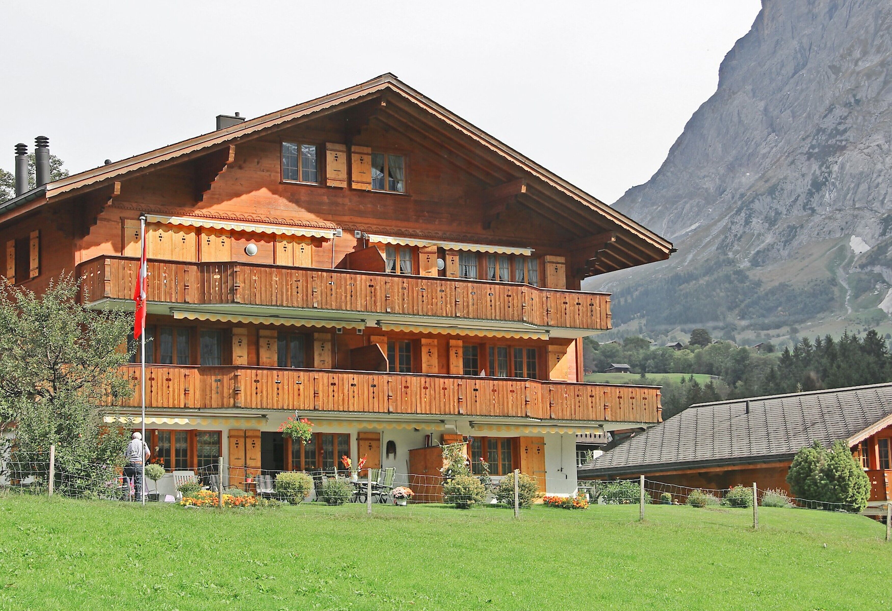 A two-story wooden chalet with balconies, surrounded by a well-maintained lawn and a mountain backdrop.