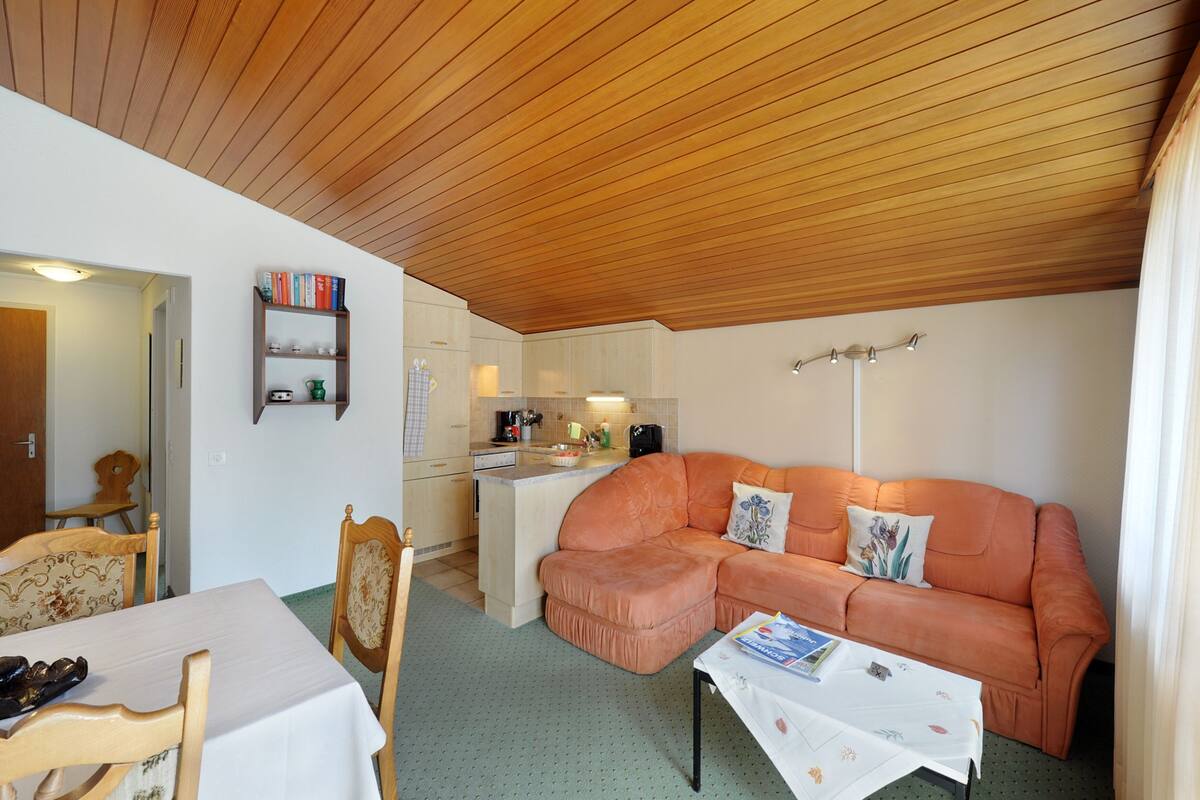 Un espacio habitable compacto con techo de madera, kitchenette, sofá, mesa de comedor y estantería.